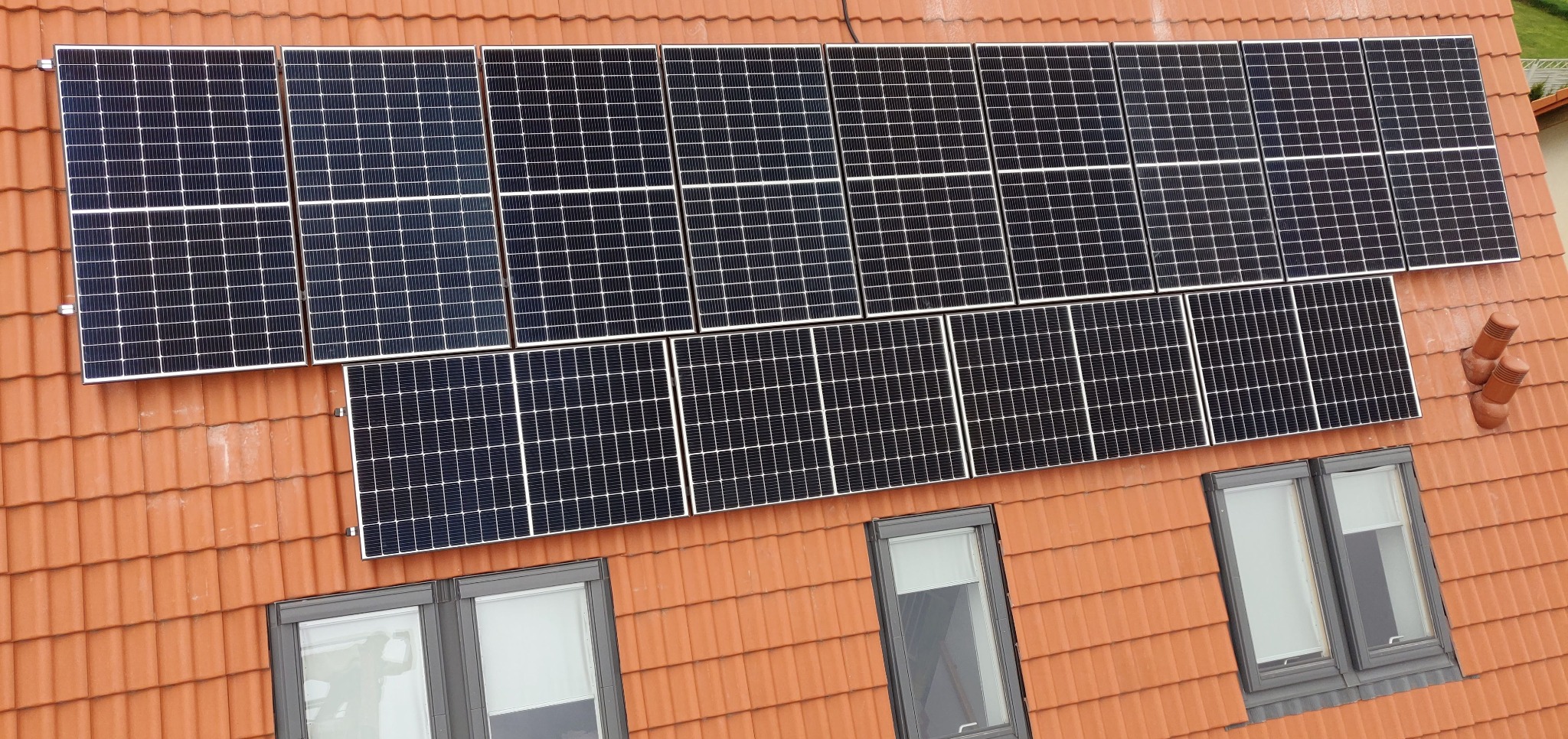 Panel słoneczny na dachu z czerwonej dachówki, widok z góry. Dwa rzędy paneli, okna dachowe w dolnej części kadru. Instalacja fotowoltaiczna na ukończonym dachu.