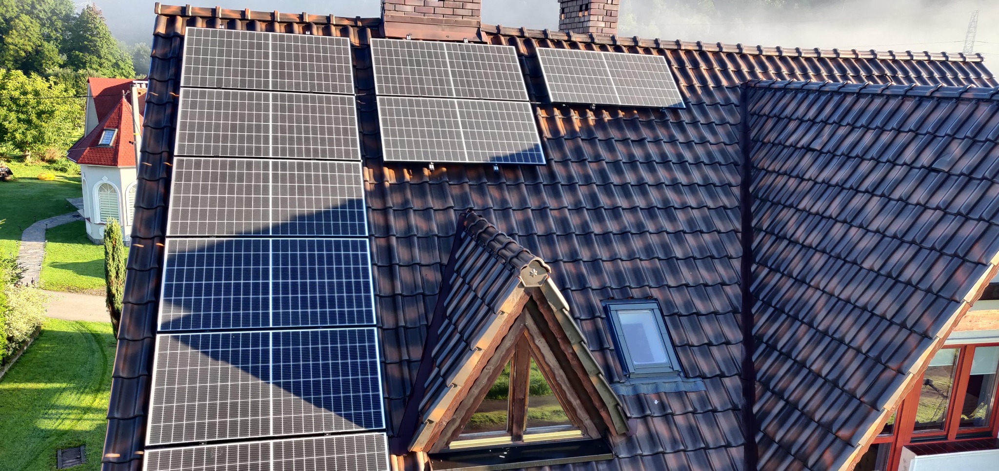 Panele fotowoltaiczne na dachu z brązowej dachówki, widok z góry, fragment okna dachowego. W tle zieleń i budynek.