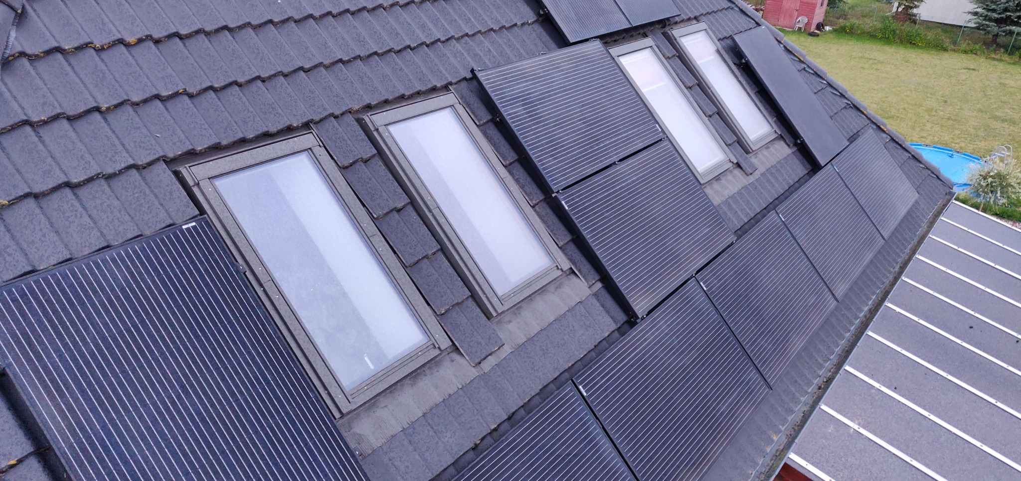 Ciemna dachówka z oknami dachowymi i panelami fotowoltaicznymi. Widok z góry na ukośny dach z zamontowanymi panelami, częściowo przysłoniętymi przez okna.