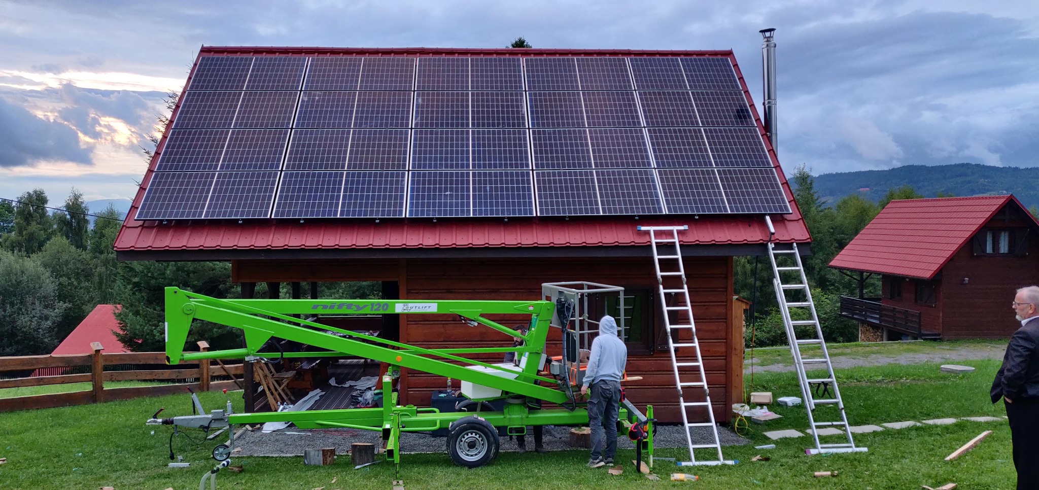 Montaż paneli słonecznych na dachu drewnianego domu z użyciem podnośnika. Widoczne drabiny i osoba w bluzie z kapturem. W tle górzysty krajobraz.