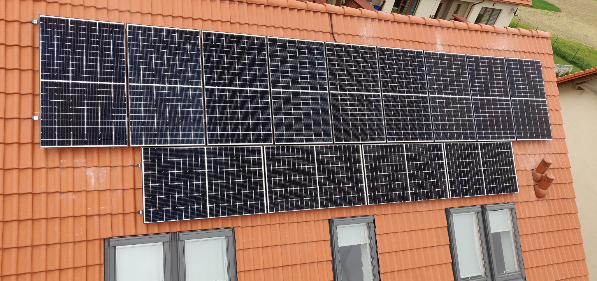 Panele fotowoltaiczne na dachu z czerwonej dachówki, widoczne okna dachowe. Układ paneli w dwóch rzędach, górny dłuższy, dolny krótszy. Ujęcie z góry, lekko z boku.
