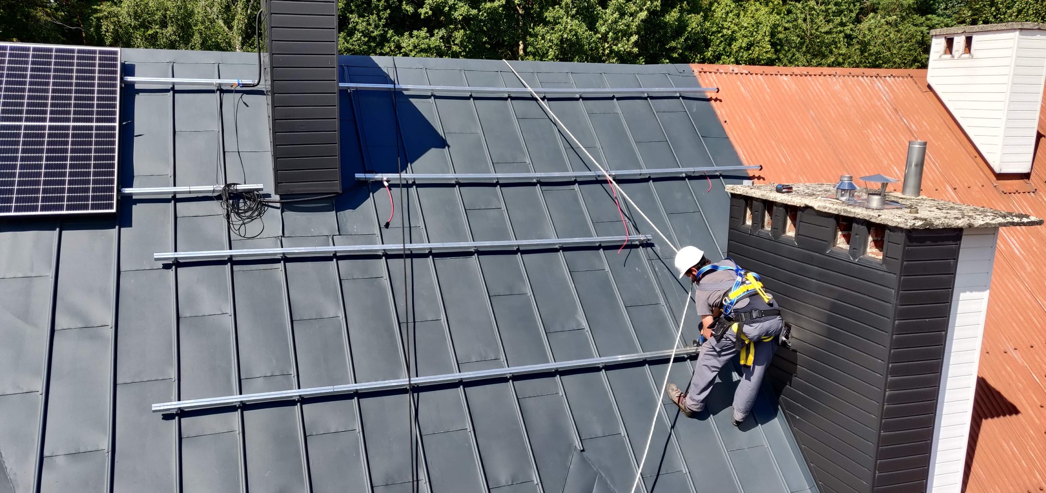 Montaż paneli słonecznych na szarym dachu z blachy. Widoczny pracownik w kasku i uprzęży, częściowo zamontowany panel i szyny montażowe. W tle komin i drzewa.