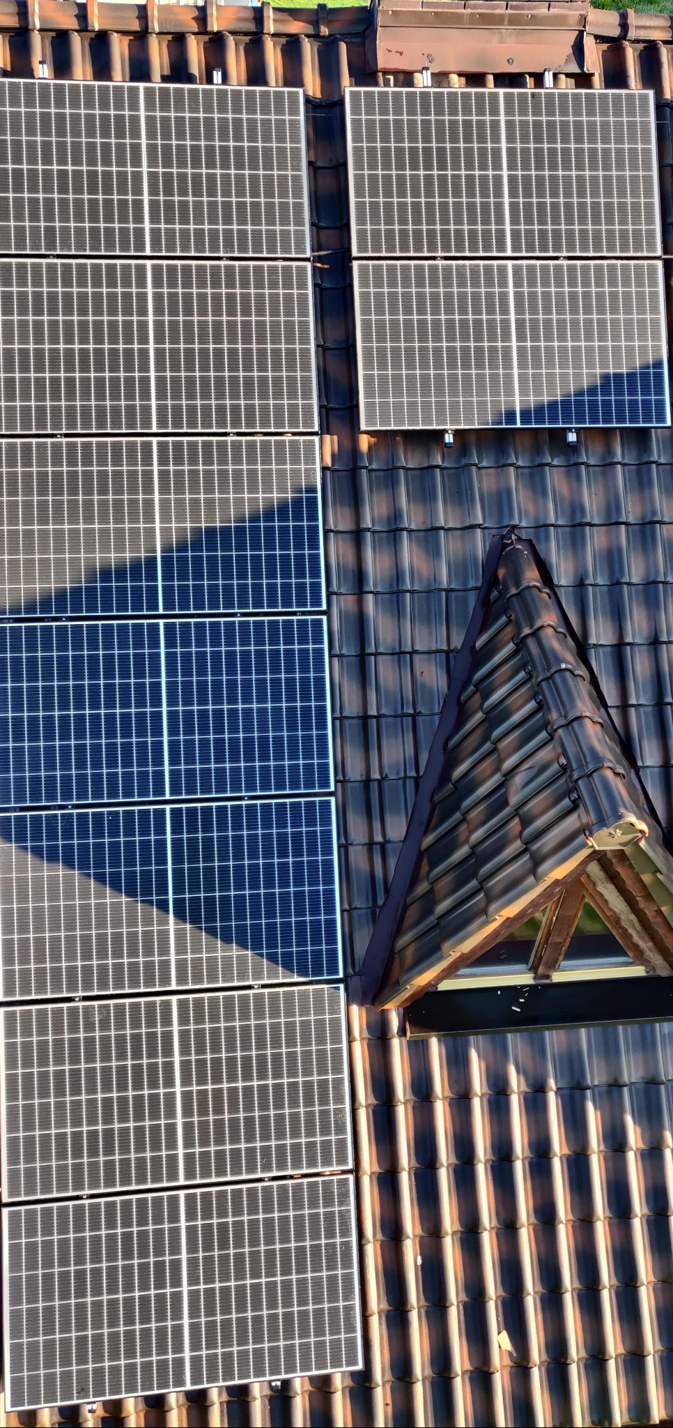 Panele fotowoltaiczne na dachu z dachówką ceramiczną i oknem dachowym. Część paneli w cieniu, widok z góry.