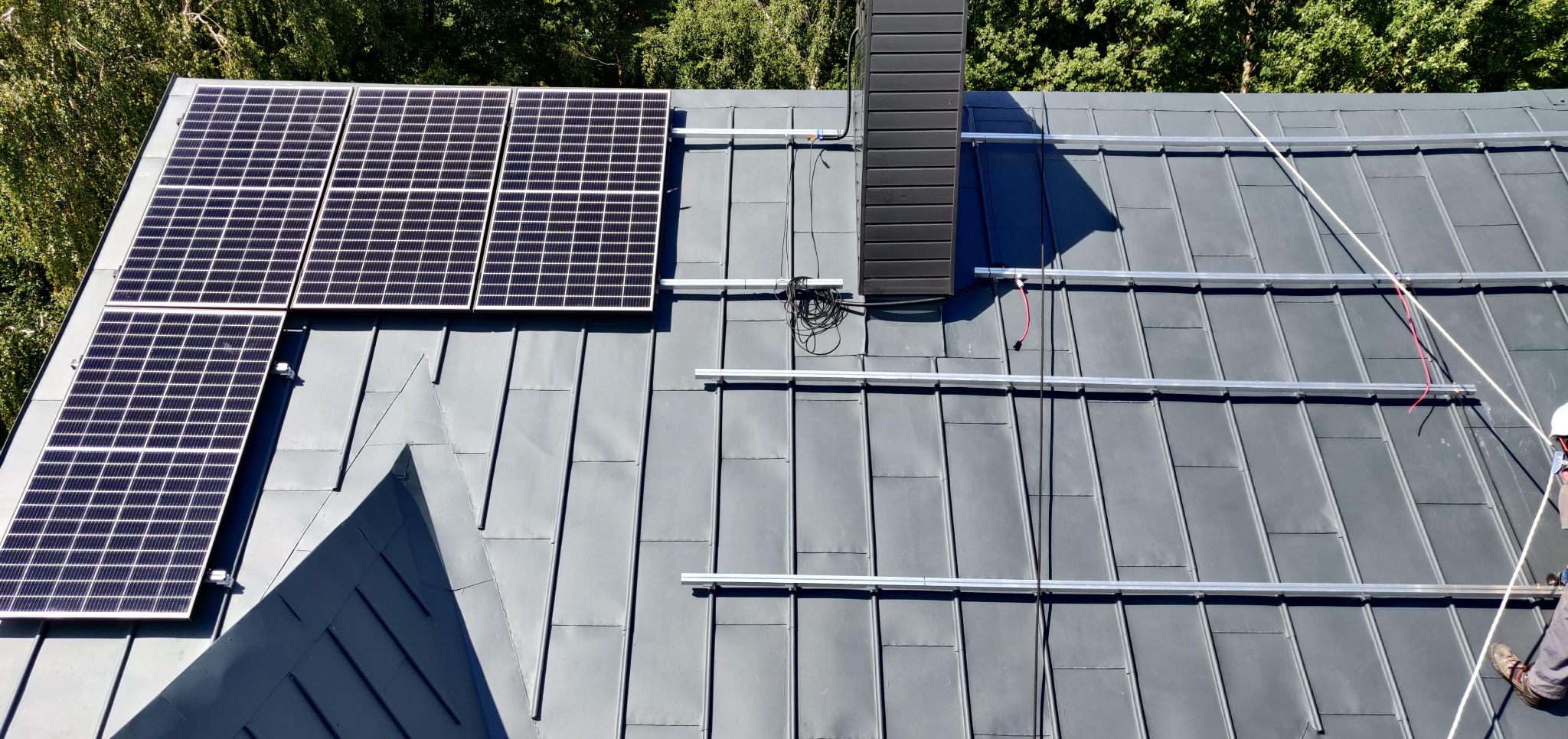 Montaż paneli fotowoltaicznych na szarym dachu blaszanym, widok z góry. Częściowo zamontowane panele i szyny montażowe. W tle zieleń drzew.