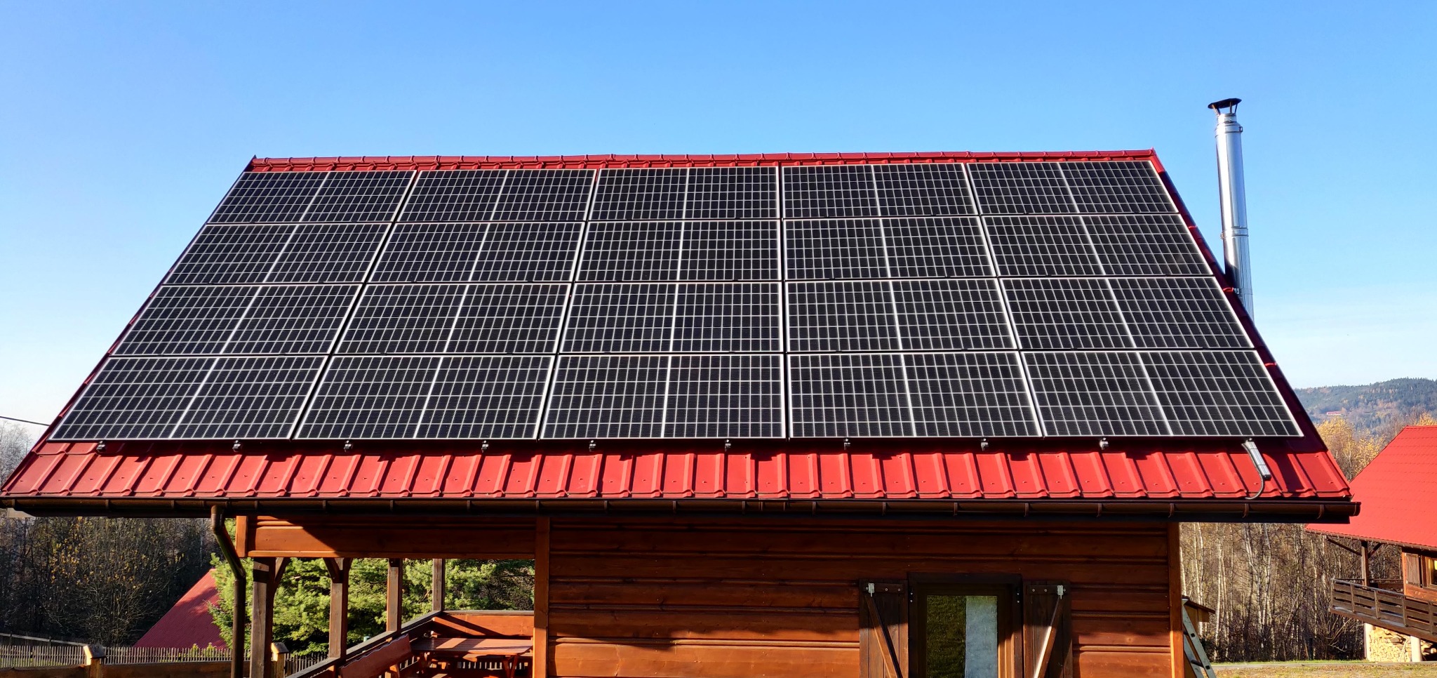 Panele słoneczne na czerwonym dachu drewnianego domu w słoneczny dzień. Widoczny komin i fragment otoczenia.