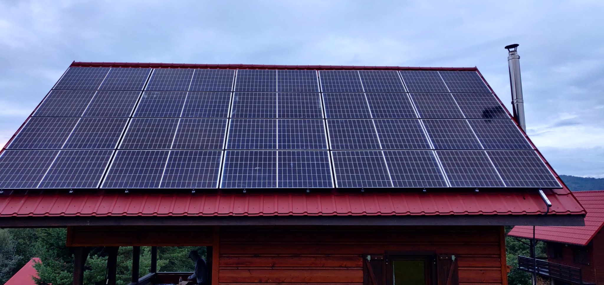 Panele słoneczne na czerwonym dachu drewnianego domu z kominem. Widok z dołu, pochmurne niebo w tle. Instalacja fotowoltaiczna na budynku.