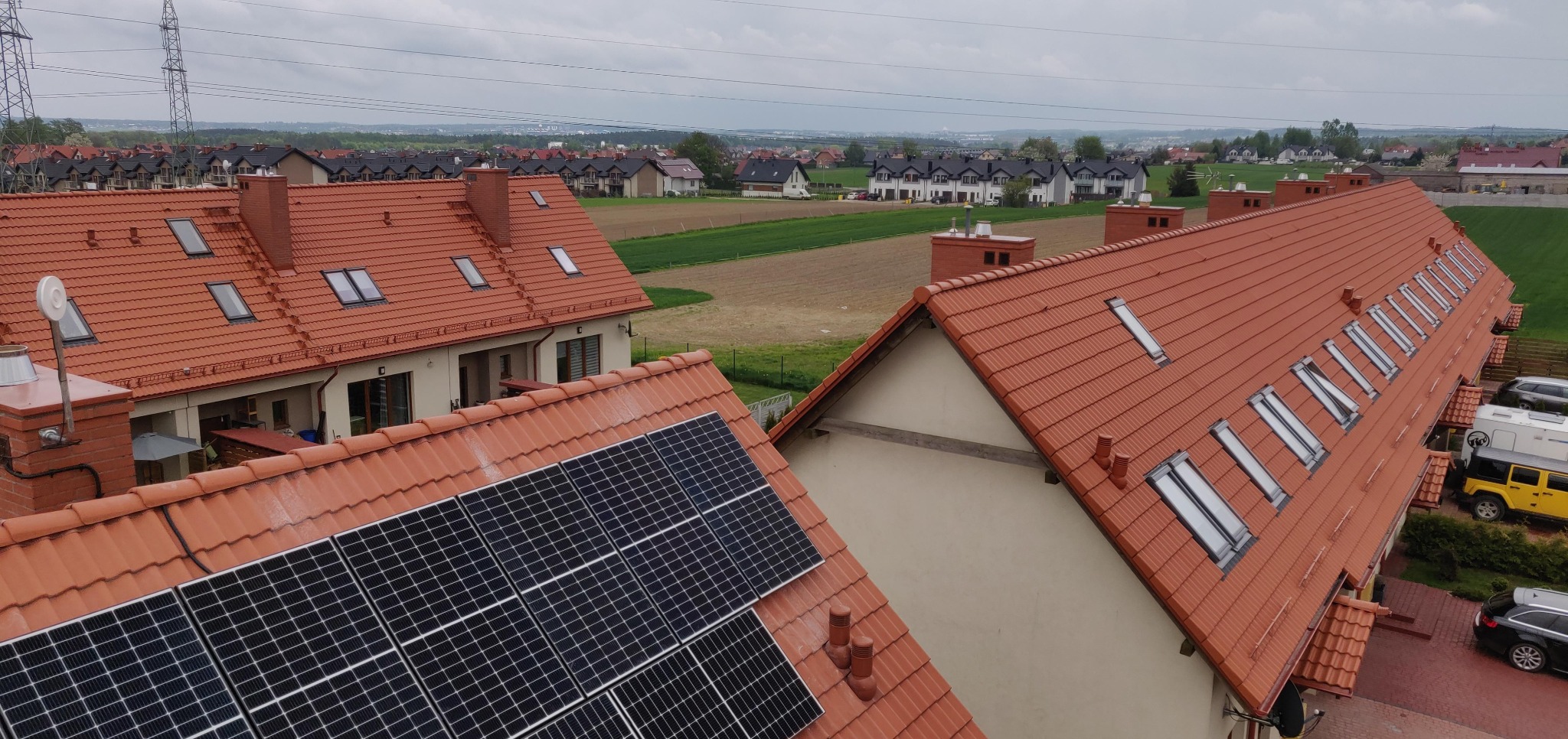 Widok z góry na domy z czerwoną dachówką i panelami słonecznymi. W tle zabudowa osiedlowa i pola uprawne pod pochmurnym niebem.