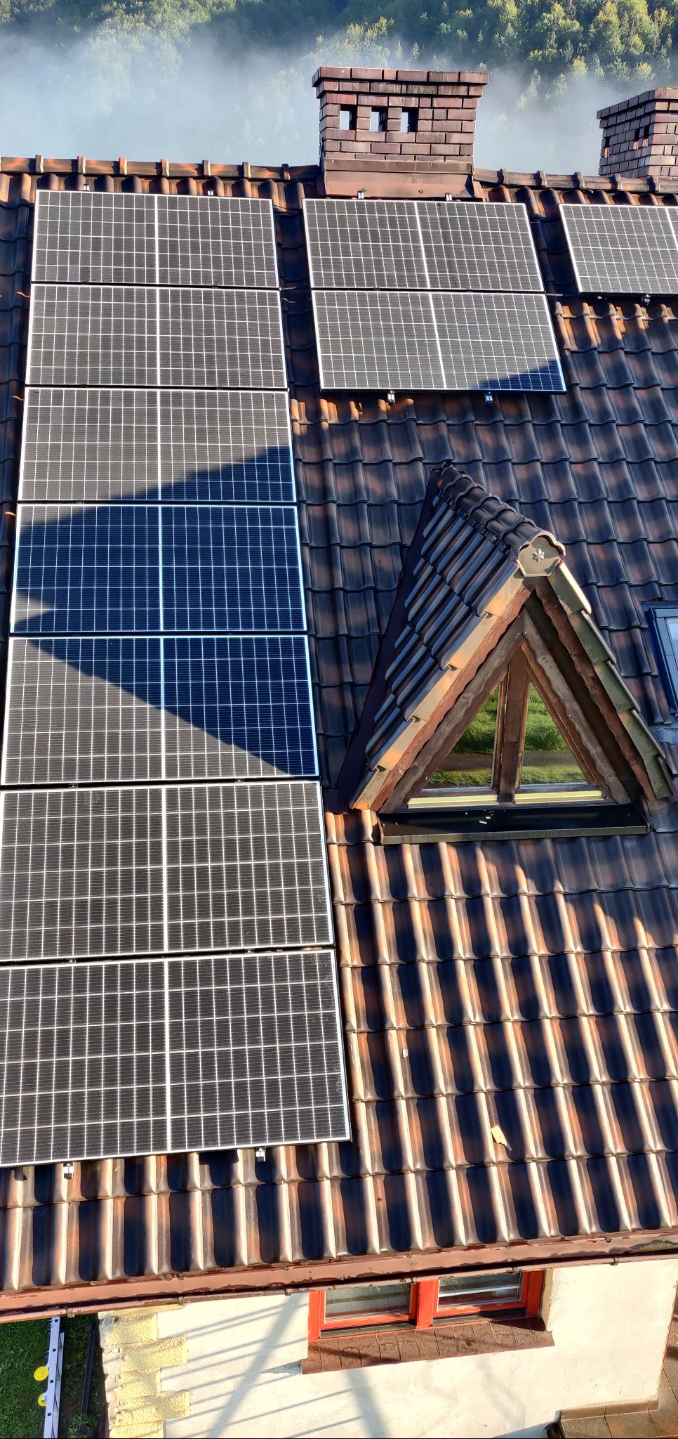Panele fotowoltaiczne na brązowym dachu z dachówki, widok z góry, komin z cegły w tle, okno dachowe trójkątne, zielone drzewa w oddali.