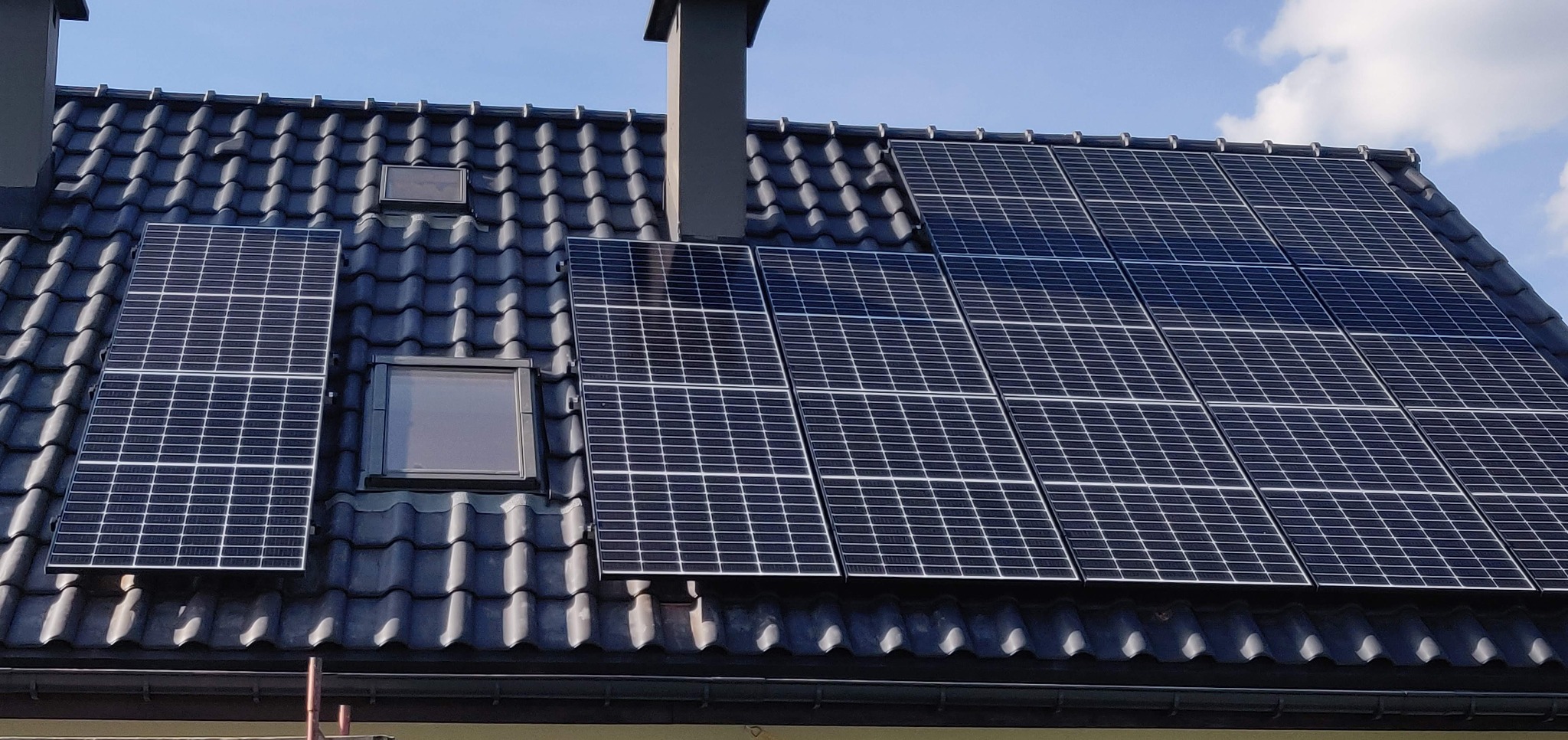 Panele fotowoltaiczne na dachu z dachówki ceramicznej, widoczne okna dachowe i komin. Ujęcie z dołu, błękitne niebo w tle. Instalacja po montażu.