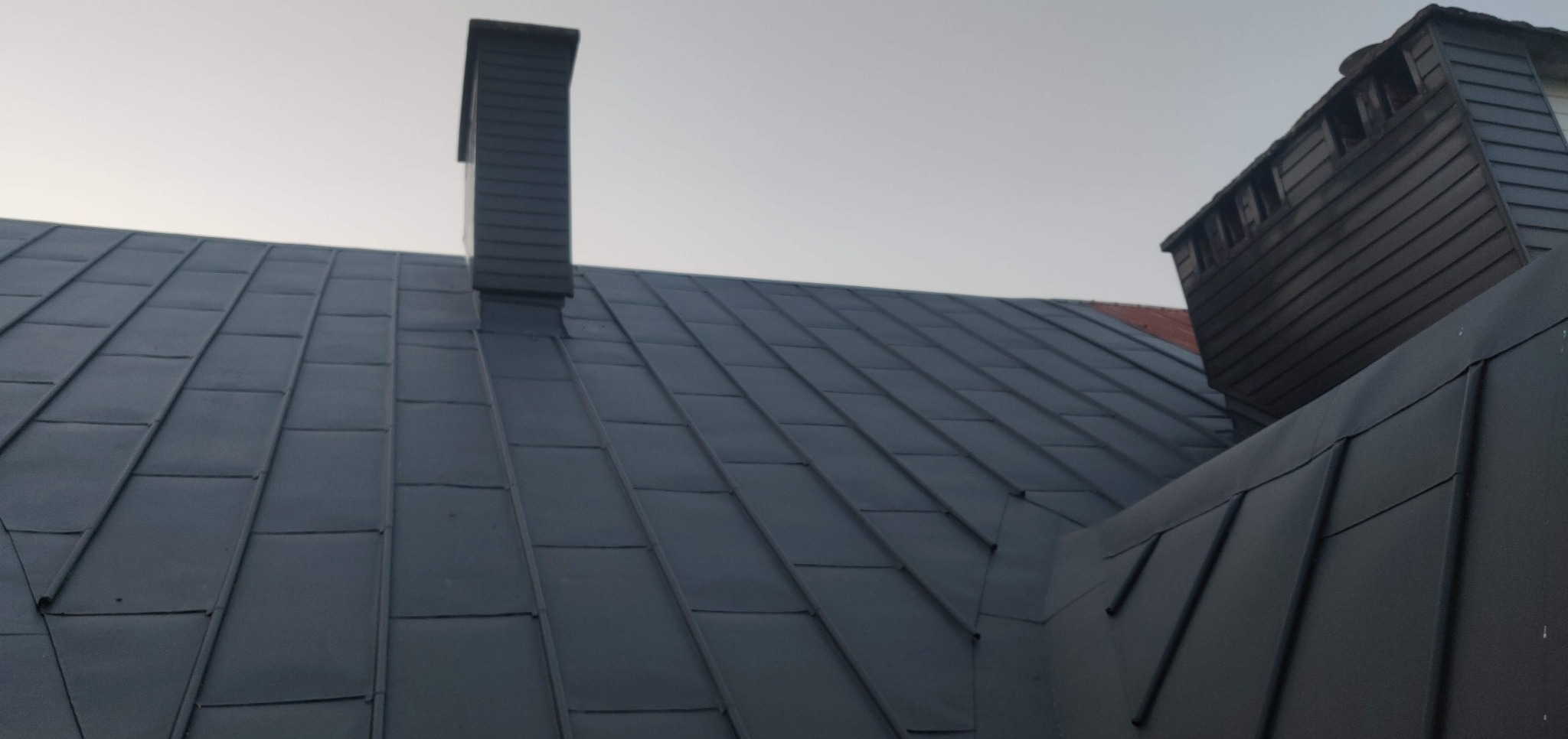 Szary dach z blachy trapezowej z dwoma kominami w odcieniach szarości, widziany pod kątem z dołu, przy pochmurnym niebie.