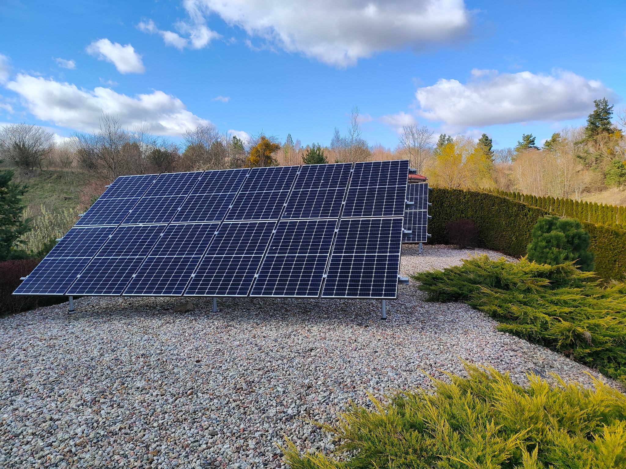 Panele fotowoltaiczne na tle błękitnego nieba z chmurami, otoczone zielenią i kamieniami. Widoczne kolejne rzędy paneli. Ujęcie z poziomu gruntu.