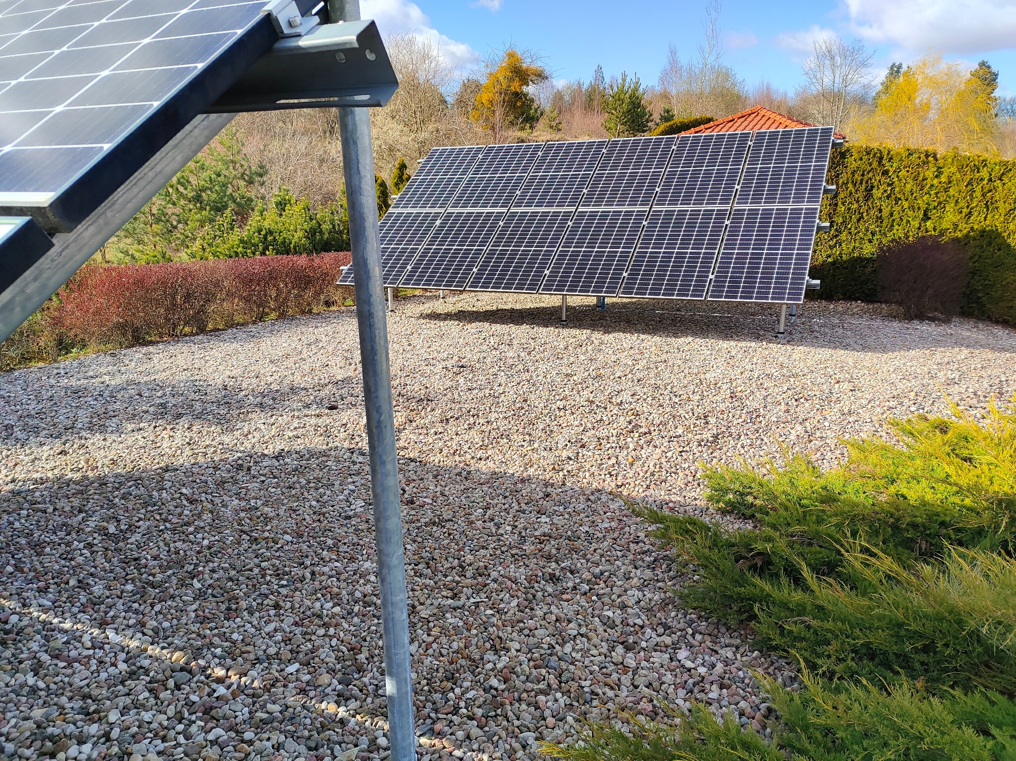 Instalacja paneli słonecznych w Przyjaźni. Dwa rzędy paneli na żwirowym podłożu, otoczone zielenią i krzewami. Błękitne niebo w tle. Widoczne mocowania i konstrukcja wsporcza.