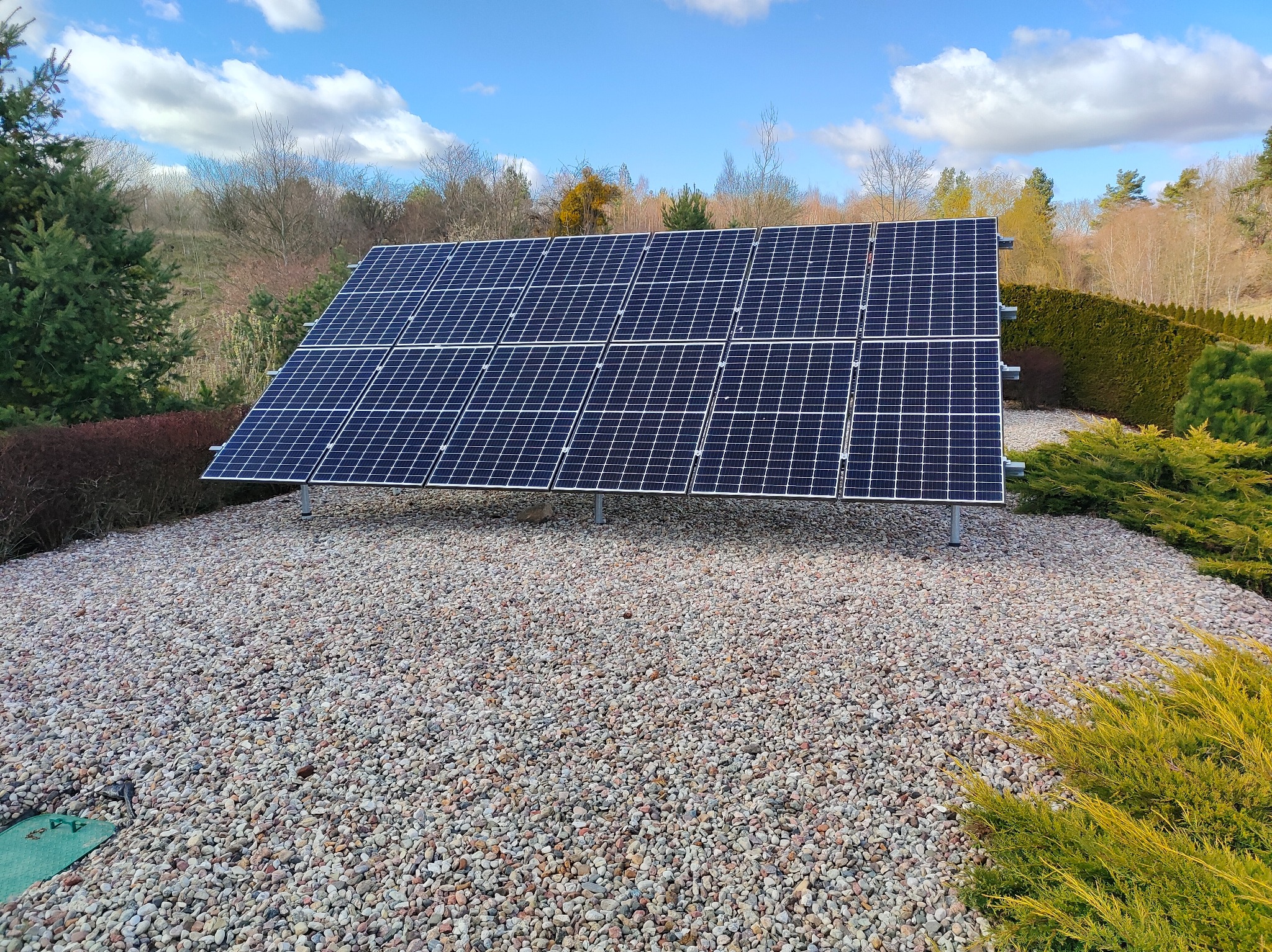 Panel fotowoltaiczny na żwirowym podłożu, otoczony zielenią, pod błękitnym niebem z chmurami. Widoczne mocowania paneli. Ujęcie z poziomu gruntu.
