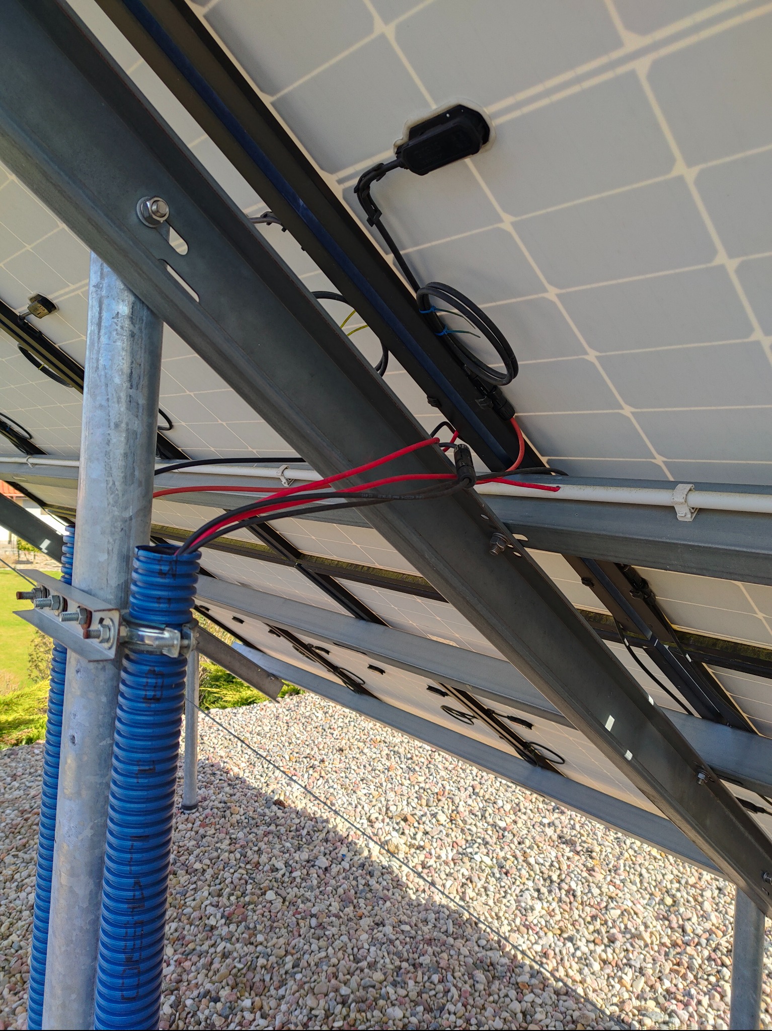 Spodnia strona paneli fotowoltaicznych z widocznymi przewodami w niebieskiej rurze karbowanej, zamocowanej do metalowej konstrukcji wsporczej.