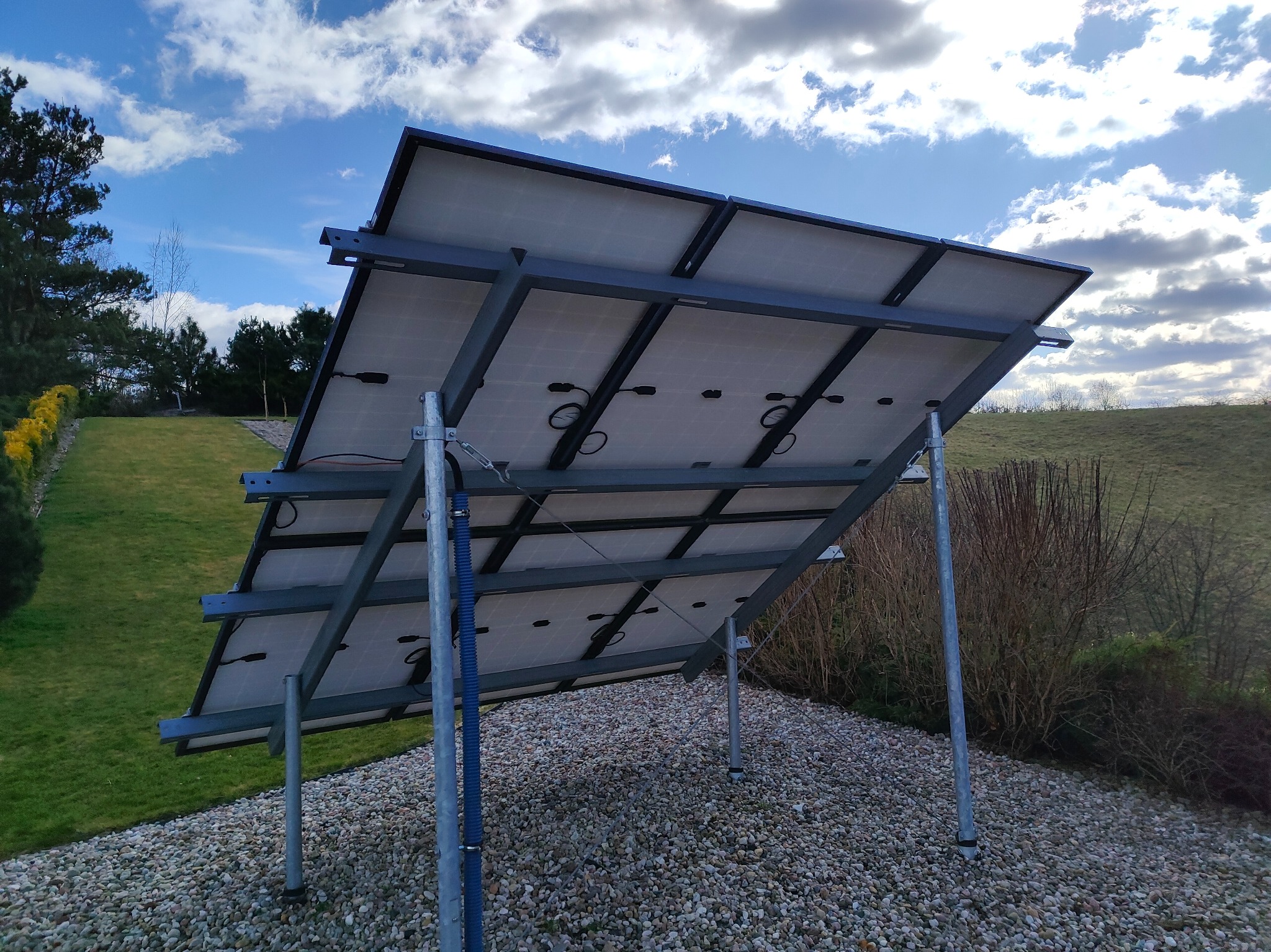 Tylny widok paneli fotowoltaicznych na metalowej konstrukcji, zamontowanych na żwirowym podłożu, z zielonym trawnikiem i drzewami w tle pod błękitnym niebem.