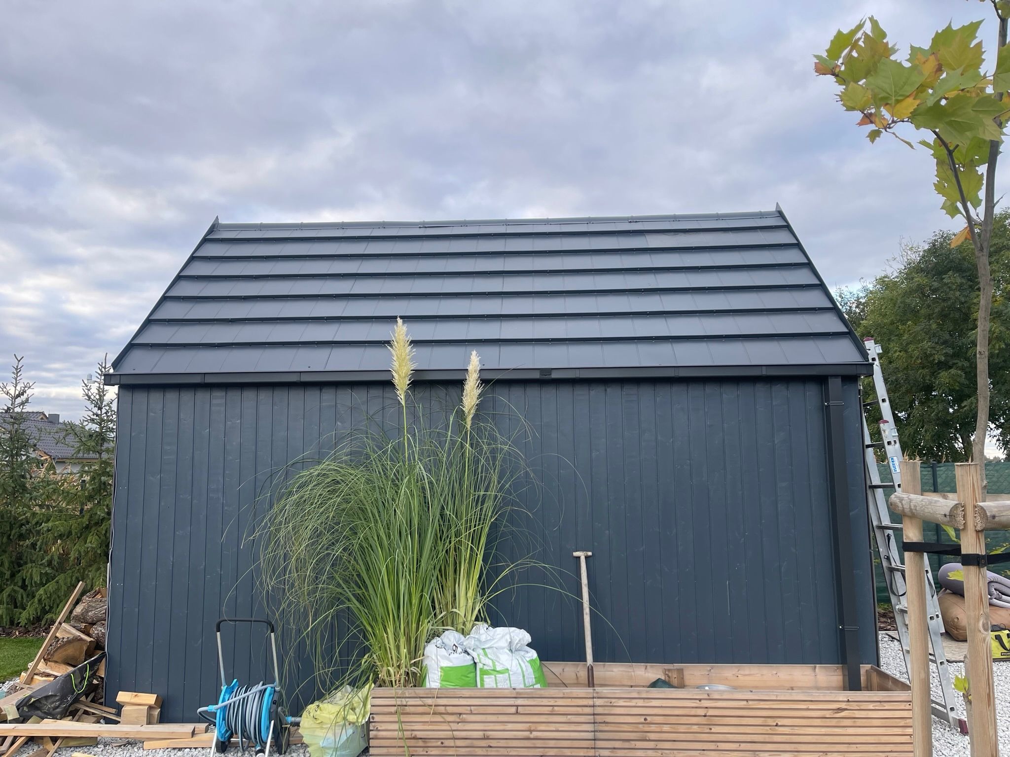 Nowoczesny, grafitowy dach z blachy na małym budynku gospodarczym, z elementami zieleni i narzędziami w tle. Ujęcie z poziomu gruntu, pochmurne niebo.