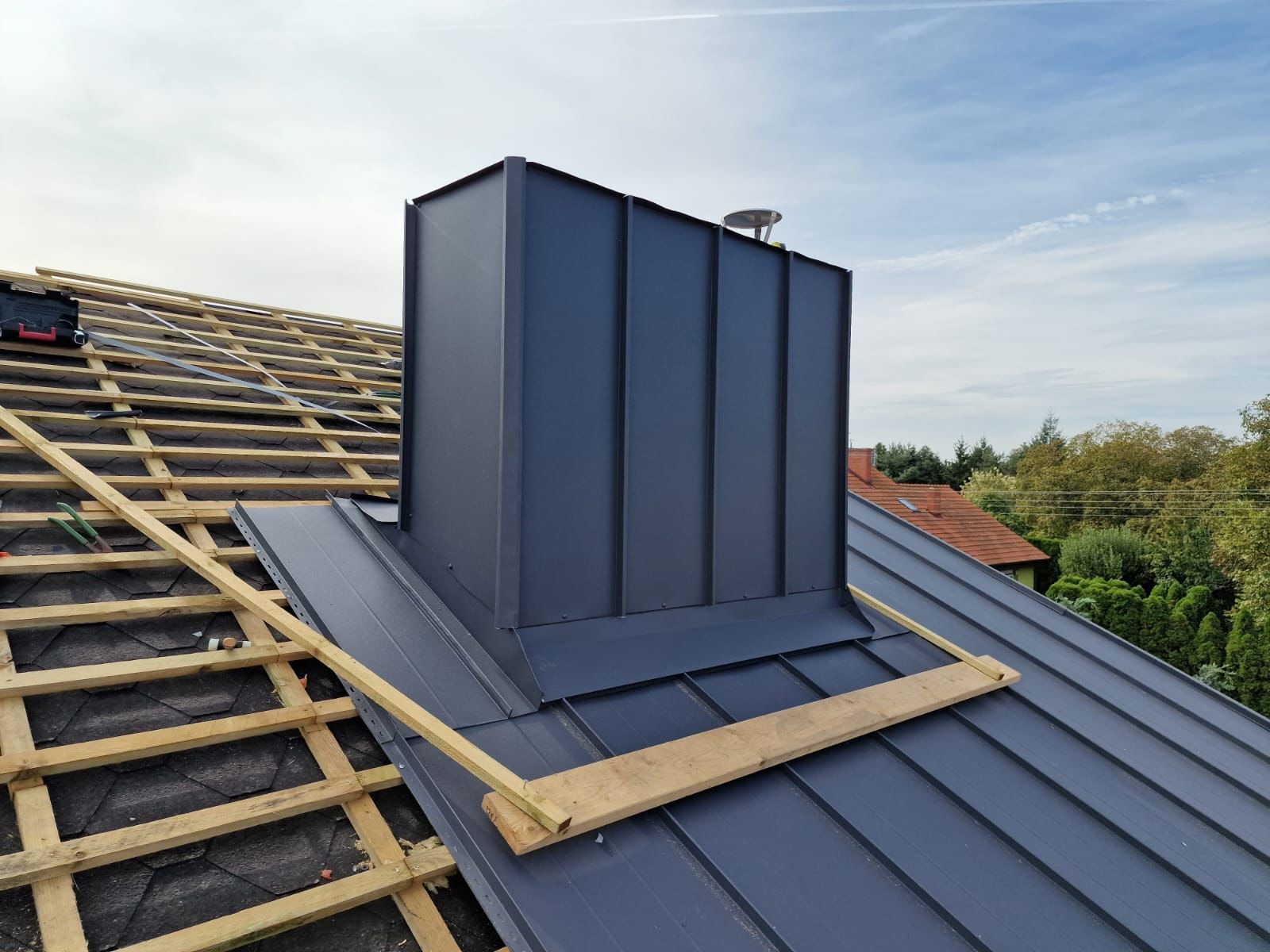 Montaż blachy na dachu z kominem w kolorze grafitowym. Widoczna więźba dachowa i elementy konstrukcyjne. Ujęcie z góry, w tle zieleń drzew i błękitne niebo.