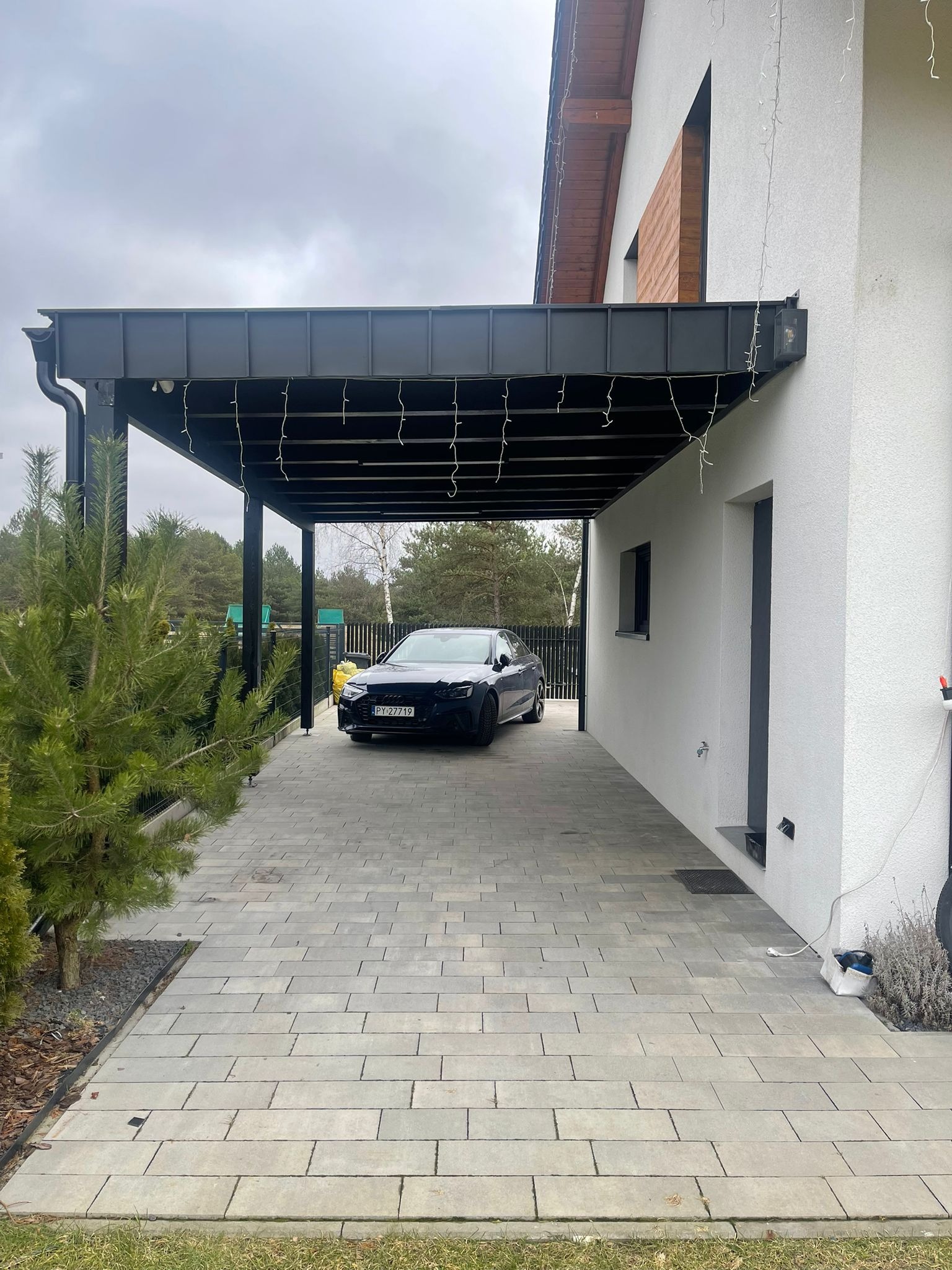 Nowoczesna wiata garażowa z czarną konstrukcją i oświetleniem, przylegająca do białego budynku. Pod wiatą zaparkowany ciemny samochód. Kostka brukowa na podjeździe.