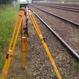 Usługi Geodezyjne Jarosław Bąk - Pomarańczowy teodolit na trójnogu ustawiony na torach kolejowych, w tle zieleń i słupy trakcji elektrycznej. Urządzenie geodezyjne w trakcie pracy.