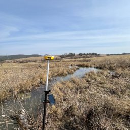 Usługi Geodezyjne Jarosław Bąk - Żółty odbiornik GNSS na tyczce z tabletem, ustawiony na zewnątrz w terenie z suchą trawą i małym zbiornikiem wodnym, w tle wzgórza i błękitne niebo.