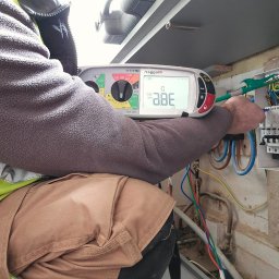 Grzegorz Stasiuk - Elektryk w trakcie pomiarów elektrycznych w rozdzielnicy, używając miernika Megger. Widoczne przewody, bezpieczniki i elementy instalacji elektrycznej.