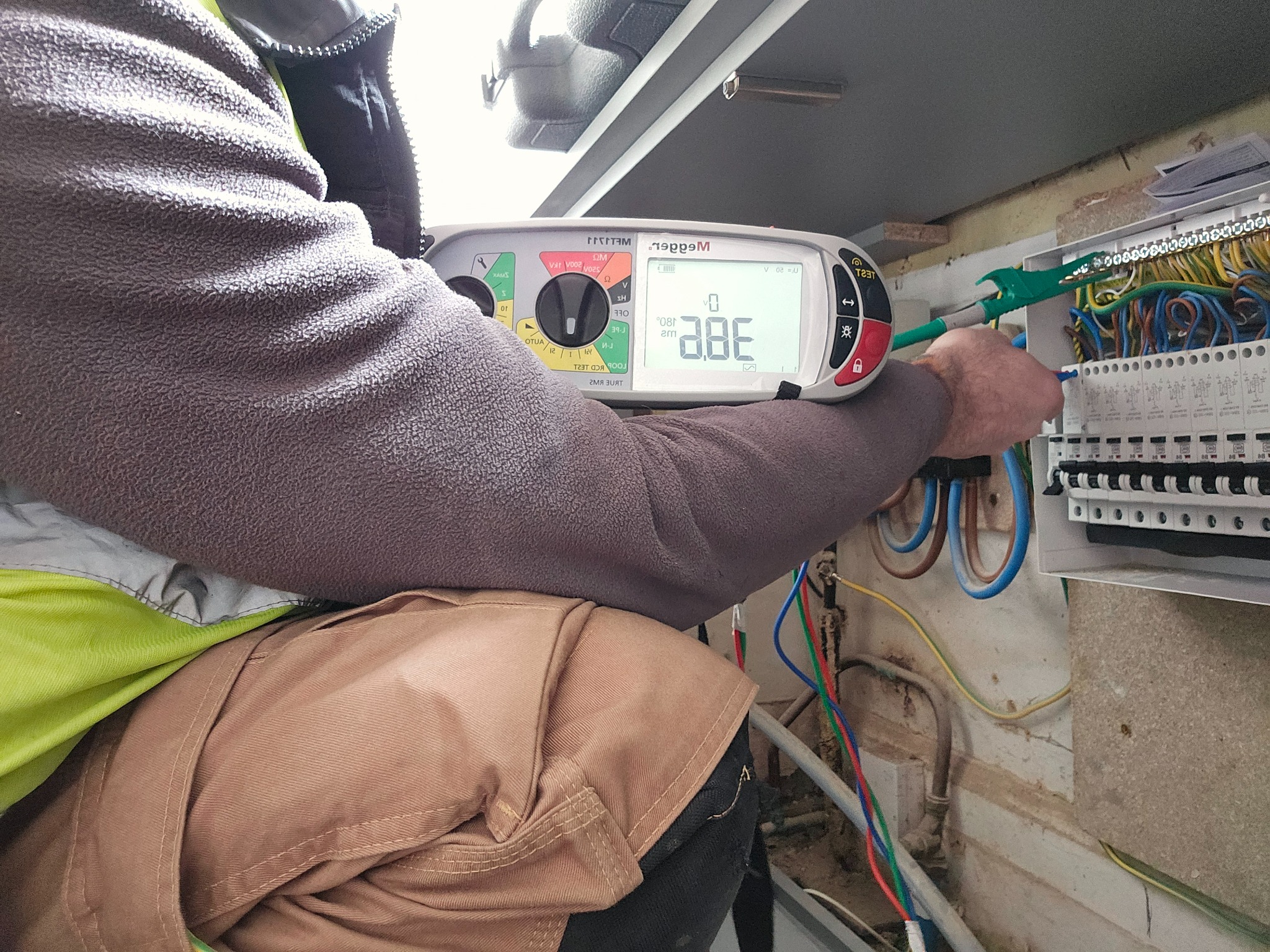 Elektryk w trakcie pomiarów elektrycznych w rozdzielnicy, używając miernika Megger. Widoczne przewody, bezpieczniki i elementy instalacji elektrycznej.