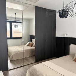 Kuźnia Mebli PRT - Nowoczesna szafa wnękowa z lustrzanymi drzwiami w sypialni. Ciemne drewno, minimalistyczny design, odbicie łóżka i okna. Stylowe oświetlenie.