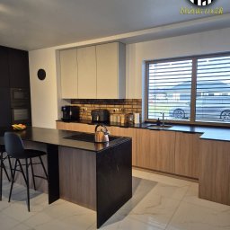 Kuźnia Mebli PRT - Nowoczesna kuchnia z wyspą: czarne blaty, drewniane fronty szafek, ceglana ściana nad blatem. Widok na dom przez okno z żaluzjami.