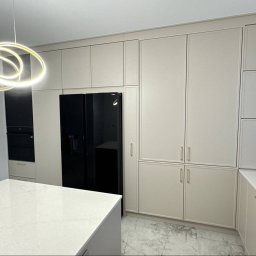 Kuźnia Mebli PRT - Beżowa zabudowa kuchenna z czarną lodówką i wyspą na marmurowej podłodze. Nad wyspą nowoczesna, okrągła lampa. Minimalistyczny design, jasne wnętrze.