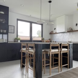 Nowoczesna kuchnia z czarnymi szafkami i wyspą z hokerami. Jasne płytki na ścianie, minimalistyczne lampy wiszące. Widok na okno i fragment blatu z dekoracjami.