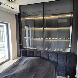 Czarna szafa wnękowa z przesuwnymi drzwiami, podświetlona, z kolekcją modeli samolotów, umieszczona nad wezgłowiem tapicerowanego łóżka w nowoczesnej sypialni.