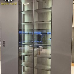 Nowoczesna szafa wnękowa z szarymi frontami i szklanymi drzwiami, podświetlona od wewnątrz. Widoczne półki i zawartość szafy. Minimalistyczny design.