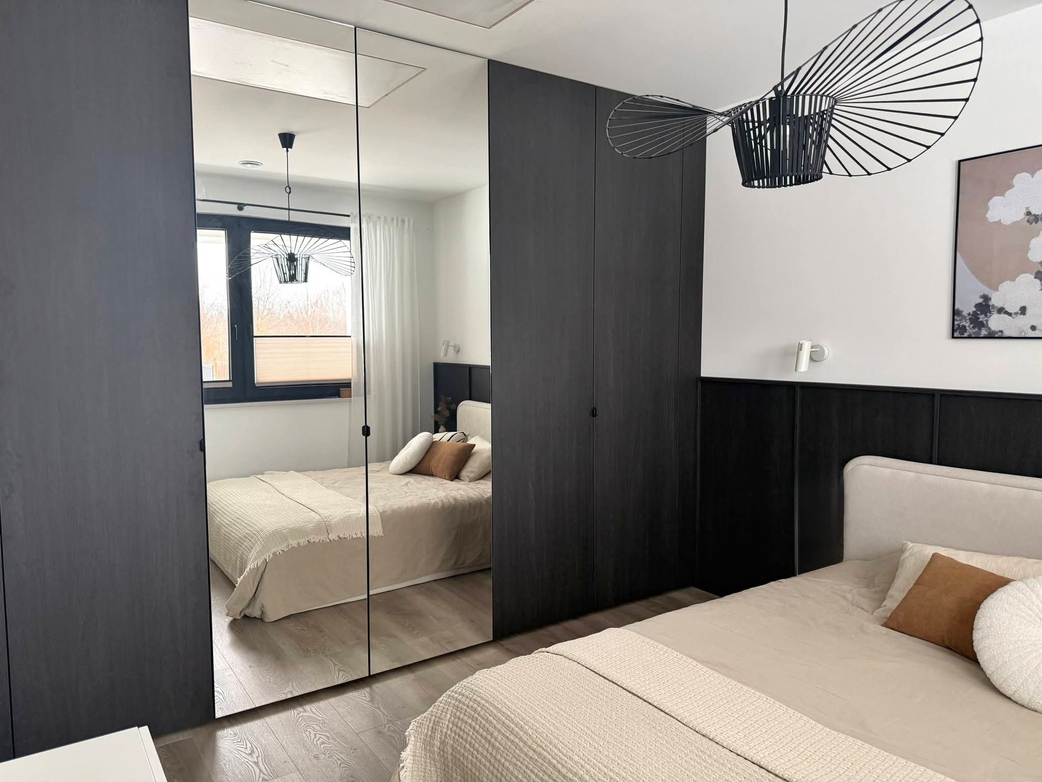 Nowoczesna szafa wnękowa z lustrzanymi drzwiami w sypialni. Ciemne drewno, minimalistyczny design, odbicie łóżka i okna. Stylowe oświetlenie.