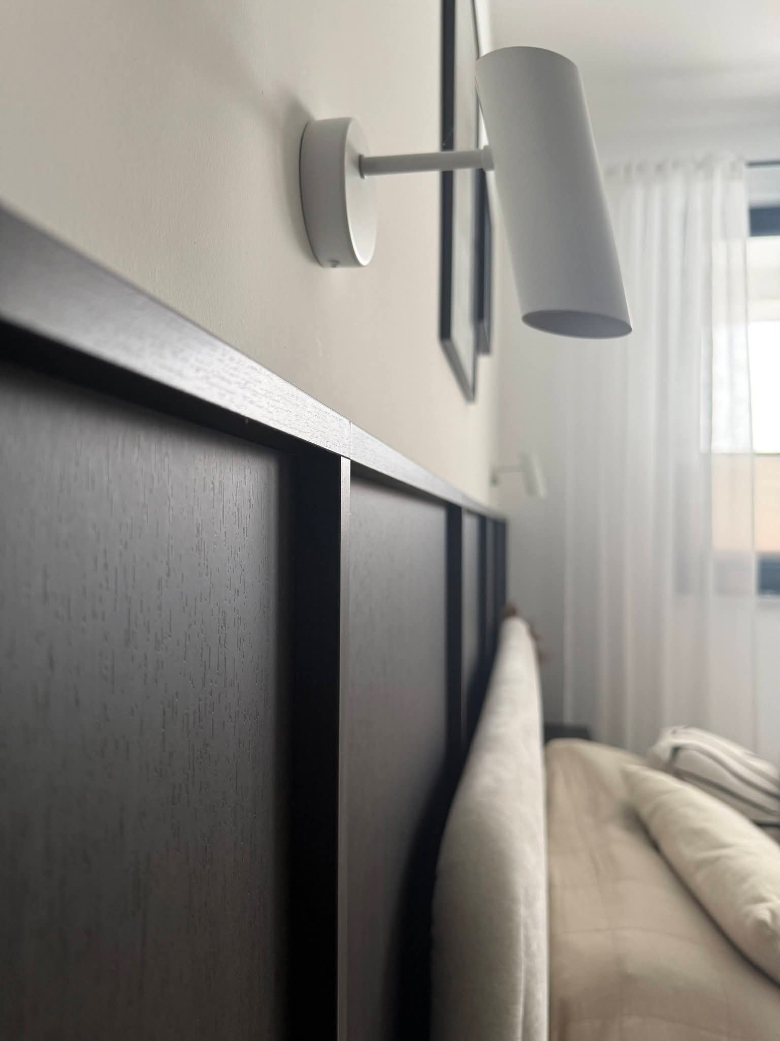 Ciemna, drewniana rama łóżka z tapicerowanym zagłówkiem, oświetlona nowoczesną lampką nocną. Minimalistyczny design sypialni z białymi zasłonami w tle.