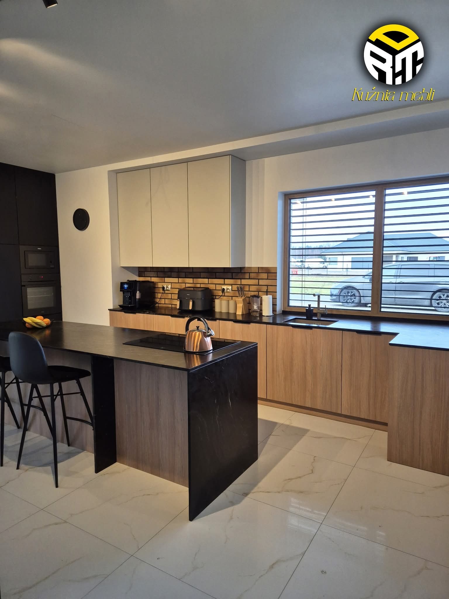 Nowoczesna kuchnia z wyspą: czarne blaty, drewniane fronty szafek, ceglana ściana nad blatem. Widok na dom przez okno z żaluzjami.