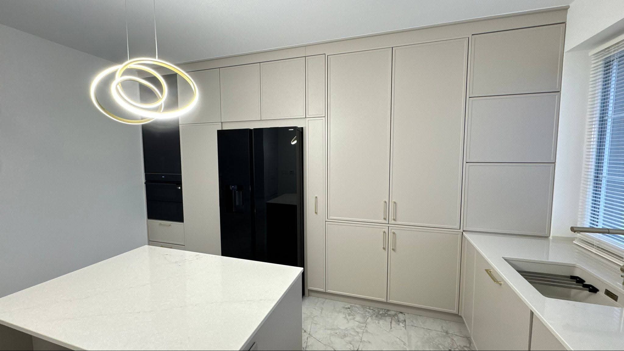 Beżowa zabudowa kuchenna z czarną lodówką i wyspą na marmurowej podłodze. Nad wyspą nowoczesna, okrągła lampa. Minimalistyczny design, jasne wnętrze.