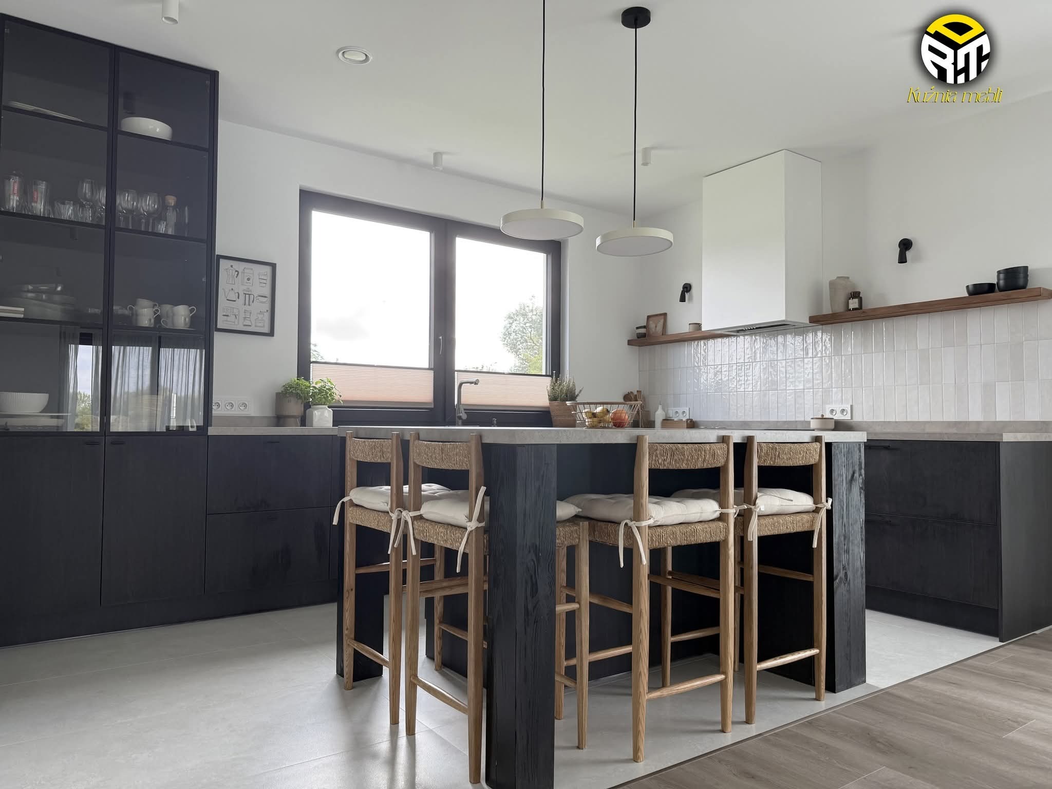 Nowoczesna kuchnia z czarnymi szafkami i wyspą z hokerami. Jasne płytki na ścianie, minimalistyczne lampy wiszące. Widok na okno i fragment blatu z dekoracjami.