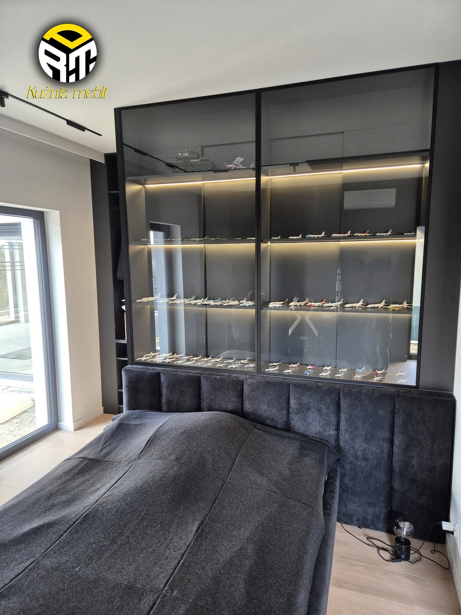 Czarna szafa wnękowa z przesuwnymi drzwiami, podświetlona, z kolekcją modeli samolotów, umieszczona nad wezgłowiem tapicerowanego łóżka w nowoczesnej sypialni.