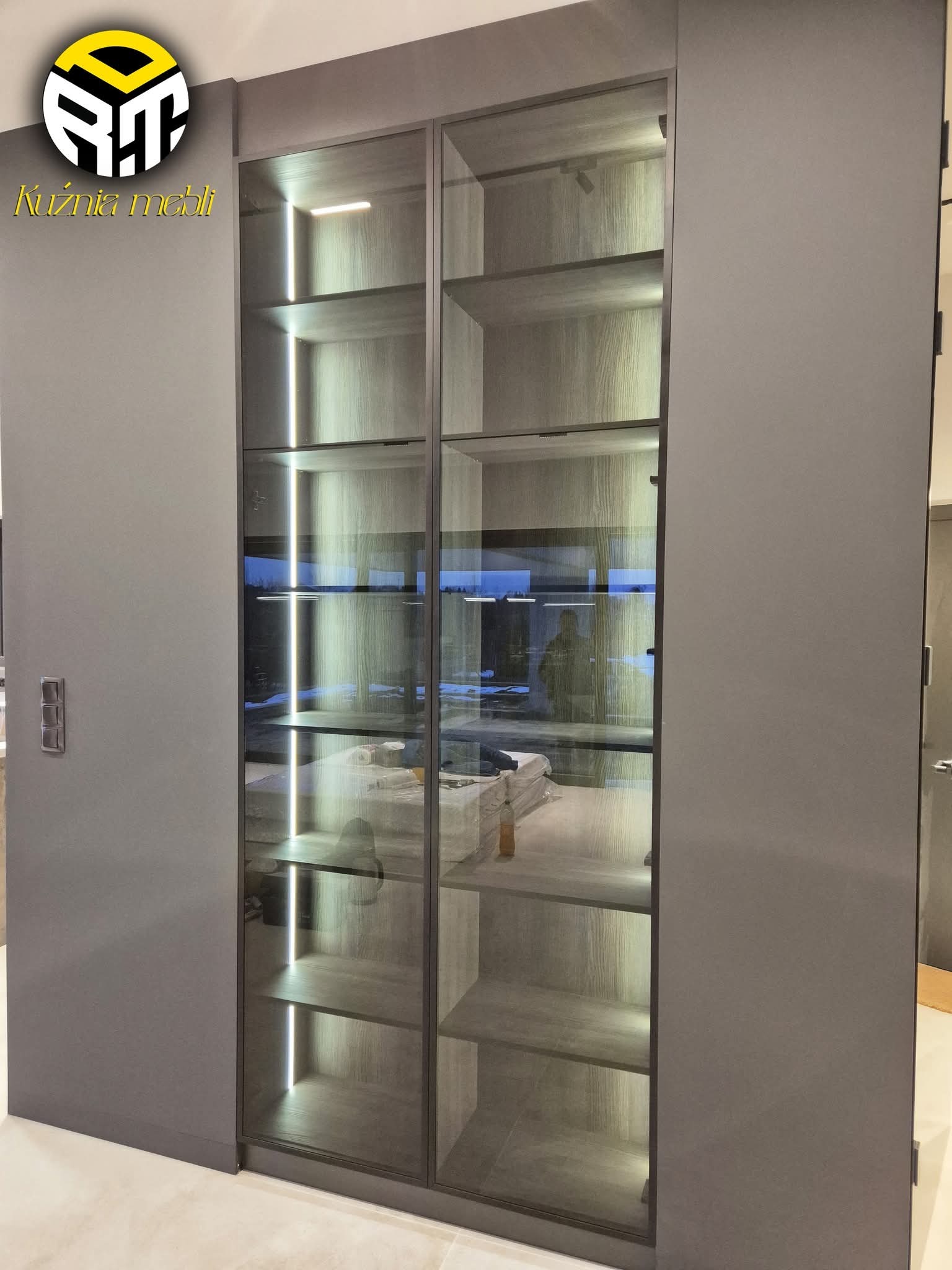 Nowoczesna szafa wnękowa z szarymi frontami i szklanymi drzwiami, podświetlona od wewnątrz. Widoczne półki i zawartość szafy. Minimalistyczny design.