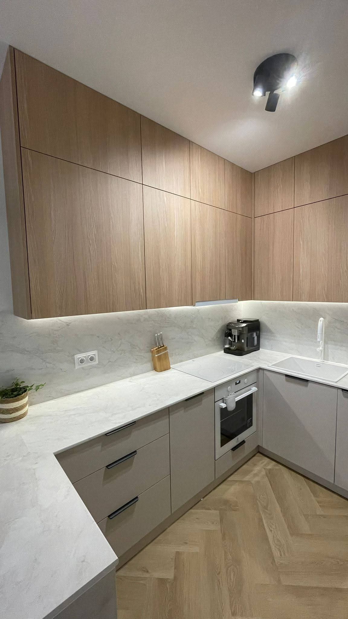 Jasna kuchnia w Tarnobrzegu z drewnianymi szafkami górnymi i szarymi dolnymi. Na blacie ekspres do kawy i deska z nożami. Parkiet w jodełkę.