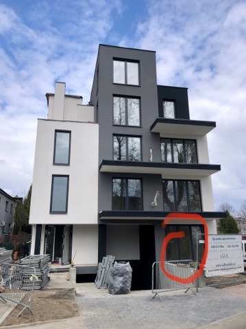 Nowoczesny budynek wielorodzinny z elewacją w odcieniach szarości i bieli, widoczne rusztowania i materiały budowlane, sugerujące prace wykończeniowe.