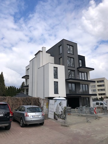 Nowoczesny budynek po renowacji elewacji, z wyraźnym podziałem na białą i szarą część. Widoczne samochody i ogrodzenie placu budowy.