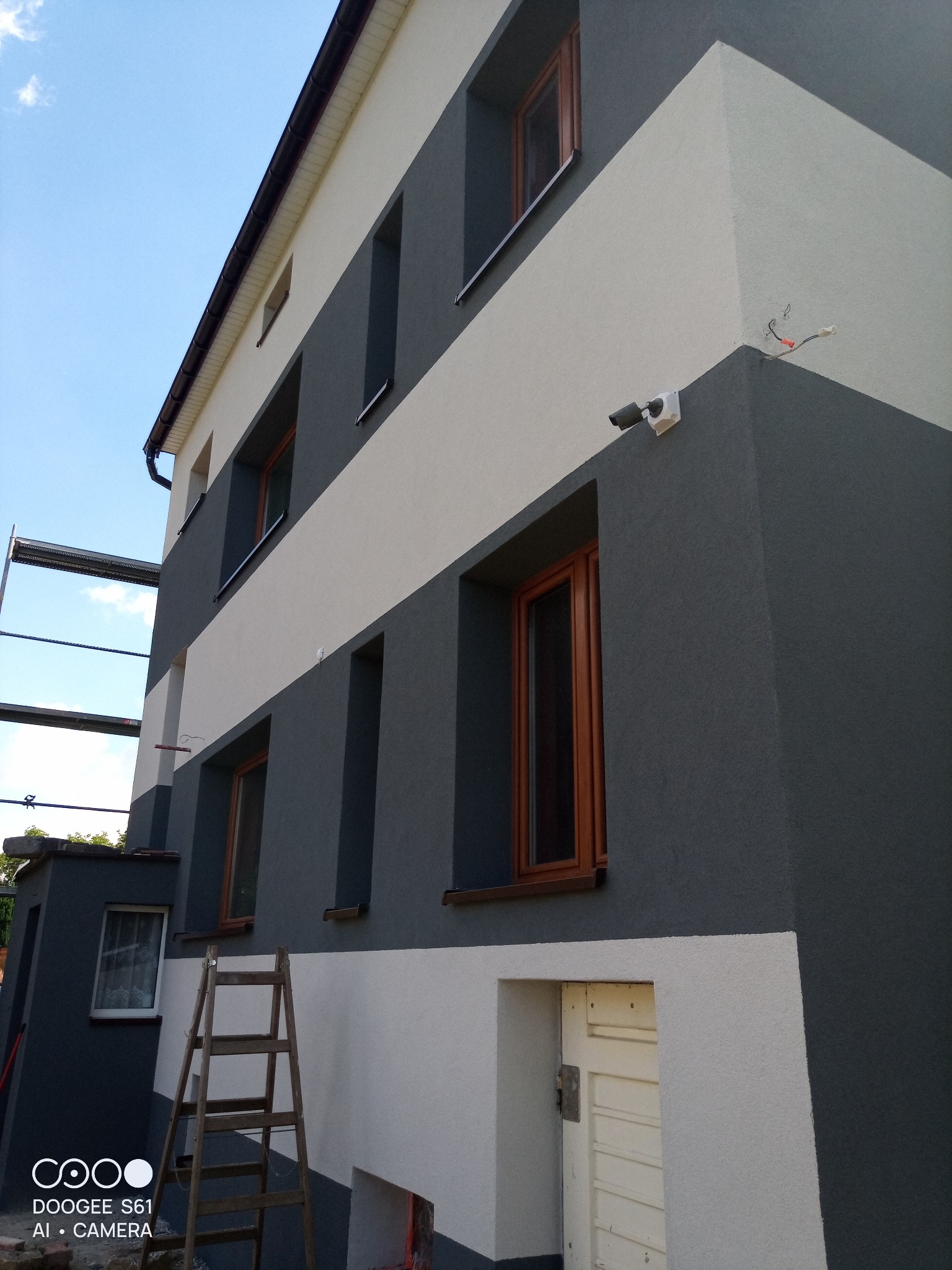 Dwukolorowa elewacja budynku po renowacji, z widocznymi oknami w drewnianych ramach i drabiną opartą o ścianę. Na rogu budynku zamontowana kamera.