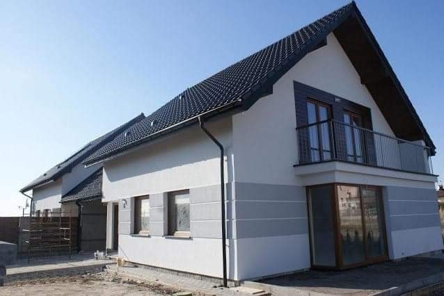 Nowoczesny dom jednorodzinny z białoszarą elewacją, ciemną dachówką i balkonem. Widoczne okna z drewnianą stolarką i fragment terenu przed budynkiem.