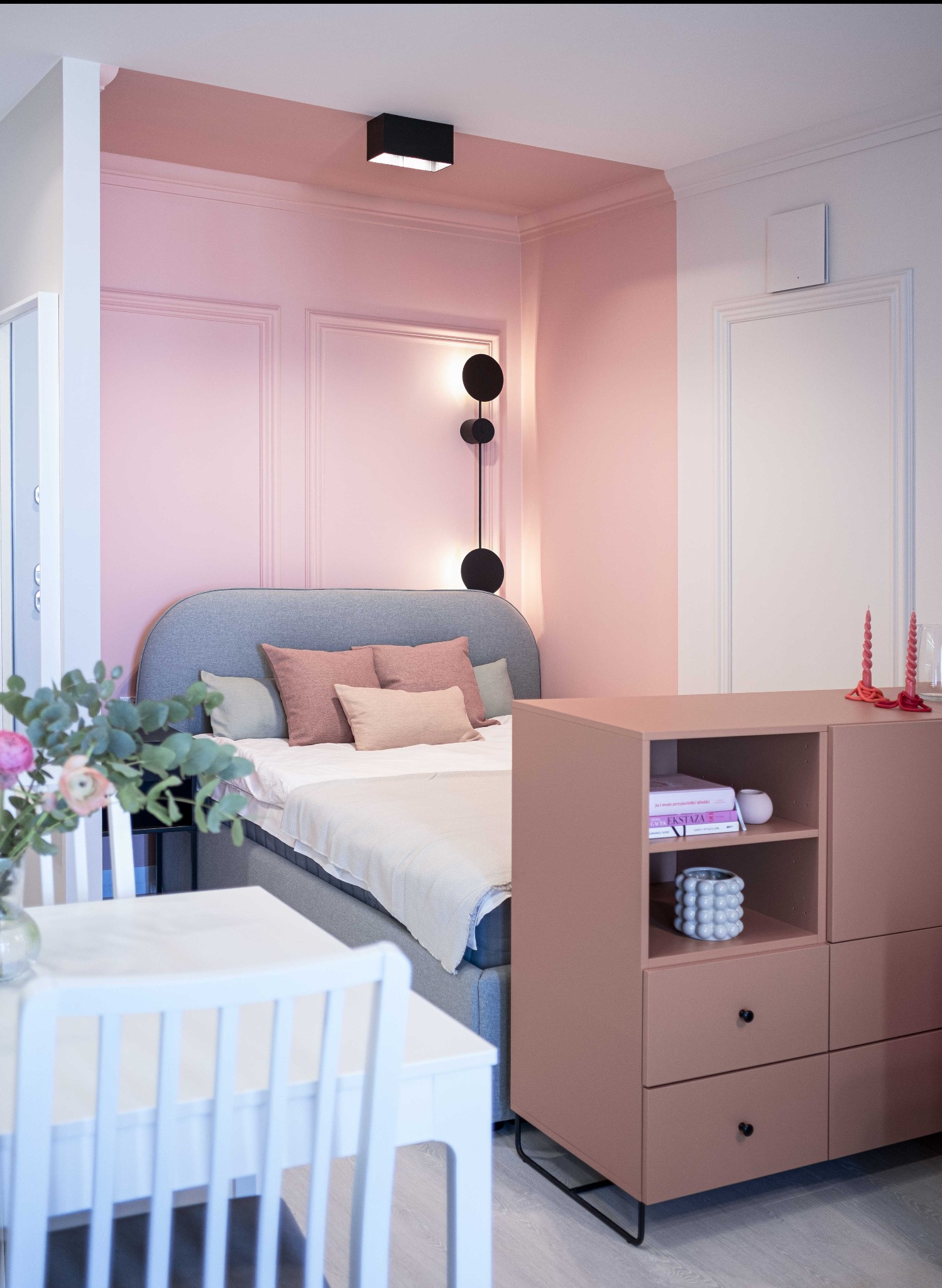 Pokój z łóżkiem w odcieniach różu i szarości, z kwiatami na stole i komodą w kolorze pudrowego różu. Nowoczesne oświetlenie i dekoracyjne panele ścienne.