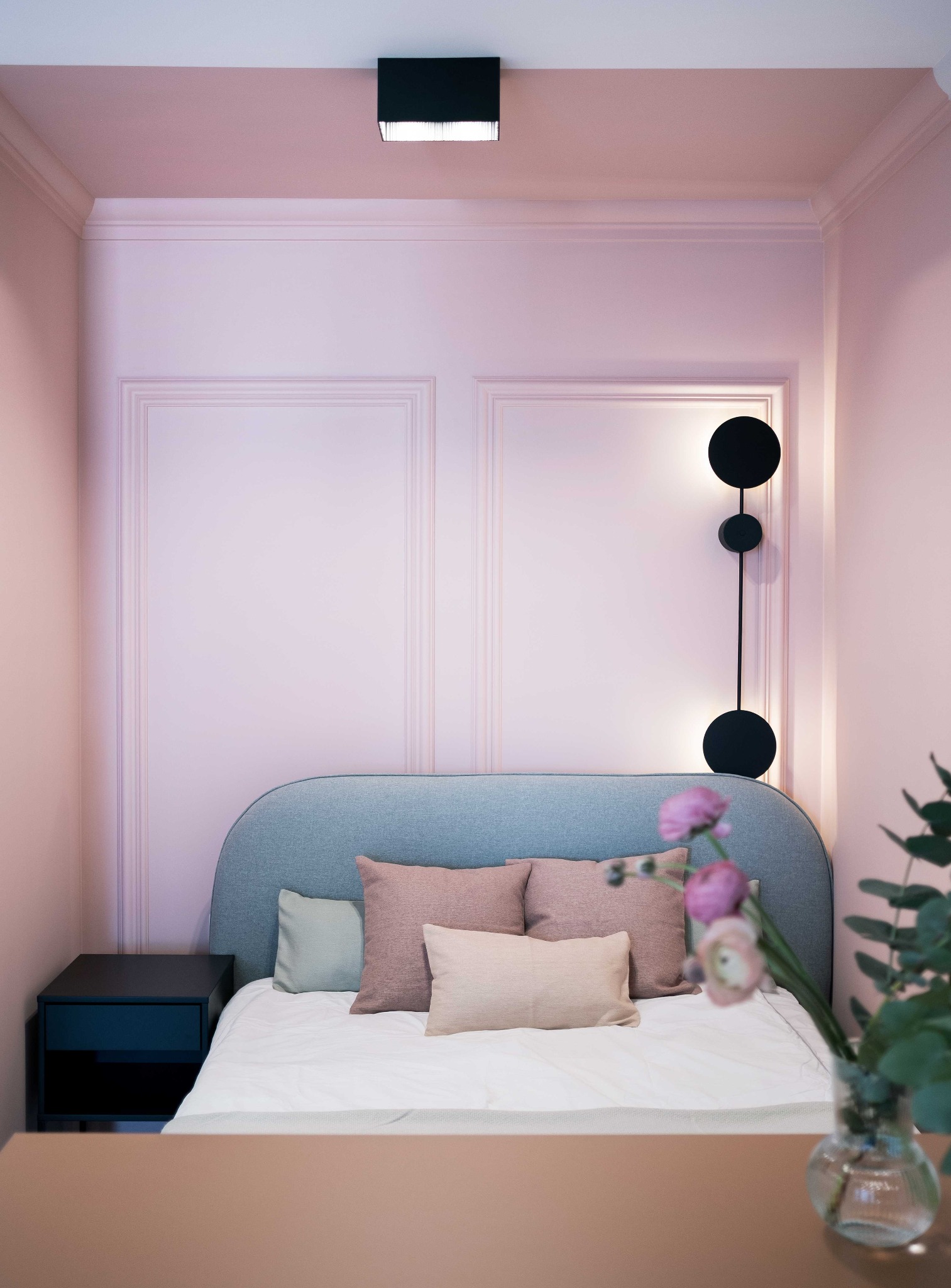 Pastelowa sypialnia z łóżkiem, kwiatami i lampą ścienną z czarnymi okręgami. Ściany w odcieniach różu z dekoracyjnymi panelami. Nowoczesny design wnętrza.