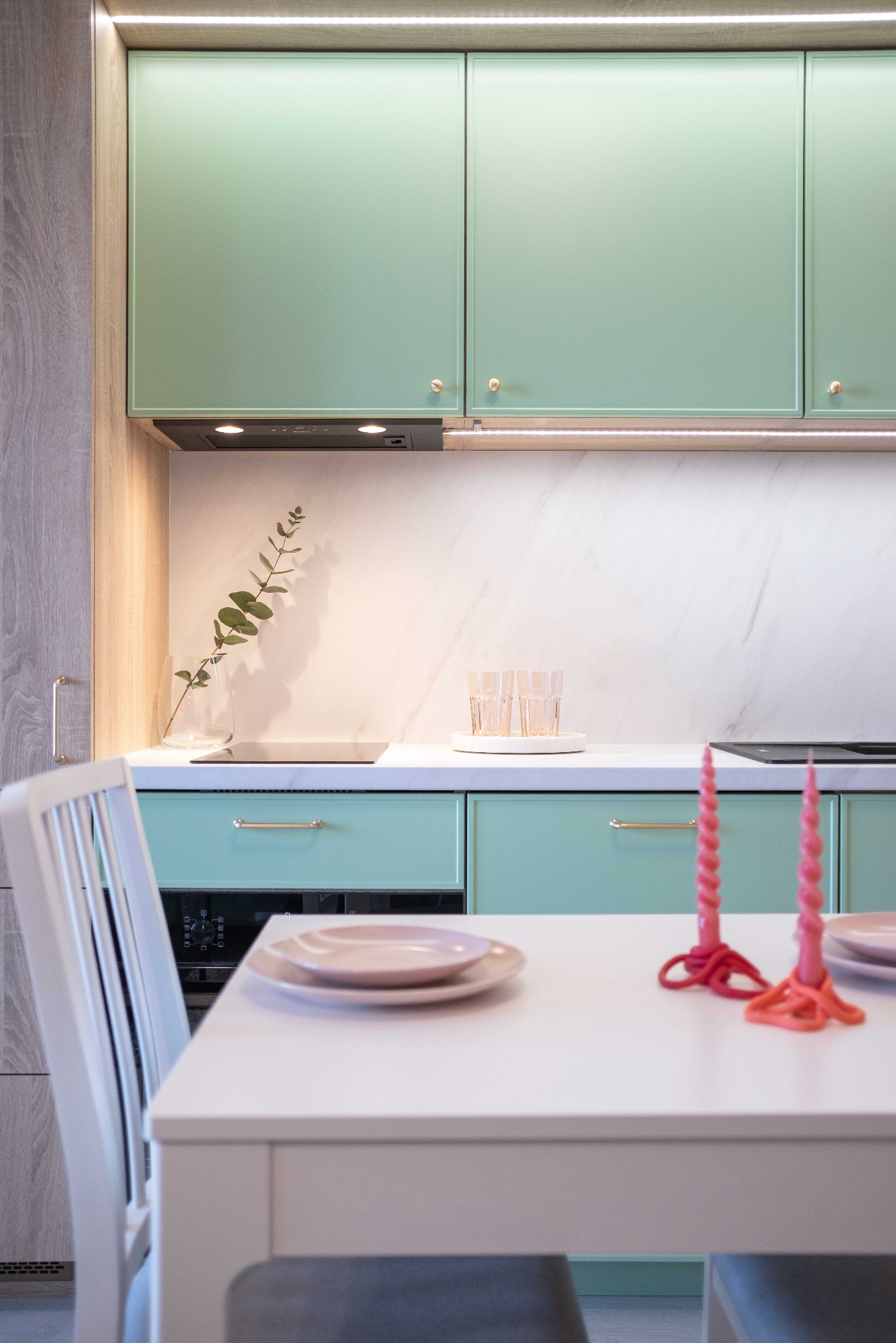 Jasna kuchnia z miętową zabudową, białym blatem i marmurową ścianą. Na stole różowe świece i talerze. Minimalistyczny design, ciepłe oświetlenie.
