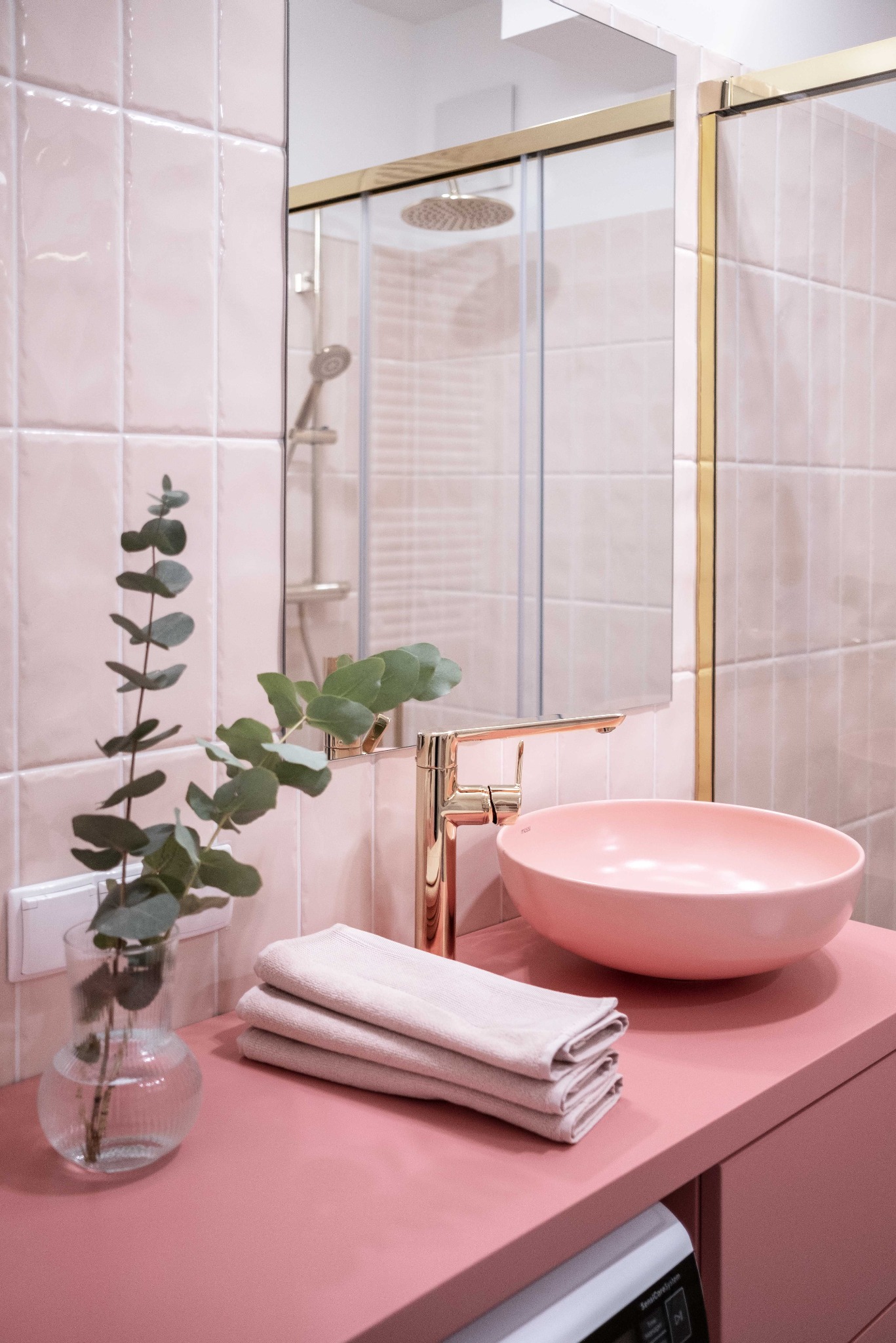 Różowa łazienka z umywalką w odcieniu pudrowego różu, złotą armaturą i eukaliptusem w wazonie. W tle kabina prysznicowa z elementami w kolorze złota.