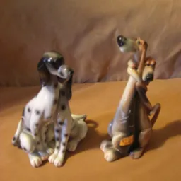 Dwie porcelanowe figurki ps&oacute;w: siedzący dalmatyńczyk z czarnymi plamkami oraz brązowo-szara figurka przedstawiająca dwa psy obejmujące się szyjami.