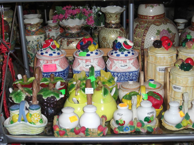 Ceramiczne pojemniki na przyprawy i cukier o różnych wzorach i kolorach, ustawione na metalowej półce. Widoczne motywy owocowe i kwiatowe, niektóre z etykietami cenowymi.